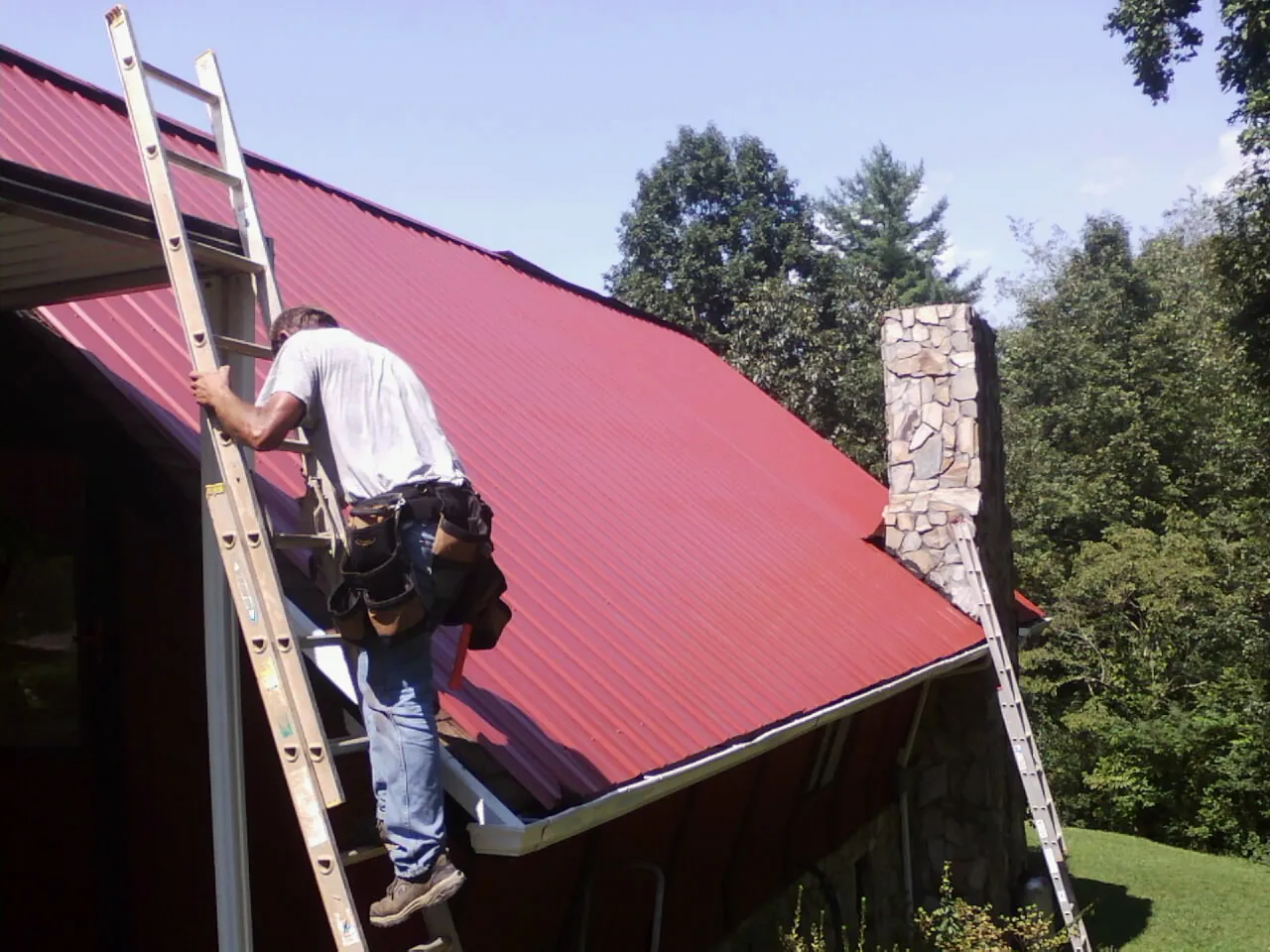 Roofer on a ladder beside a red metal roof and stone chimney — Soffit & Fascia Repair by Roofing Brothers in Saint Francis